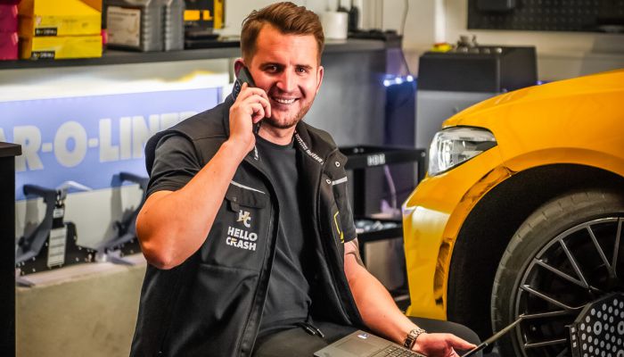 Unfallgutachter von HelloCrash bei der digitalen Fahrzeugbewertung mit Laptop in moderner Werkstatt neben beschädigtem Auto